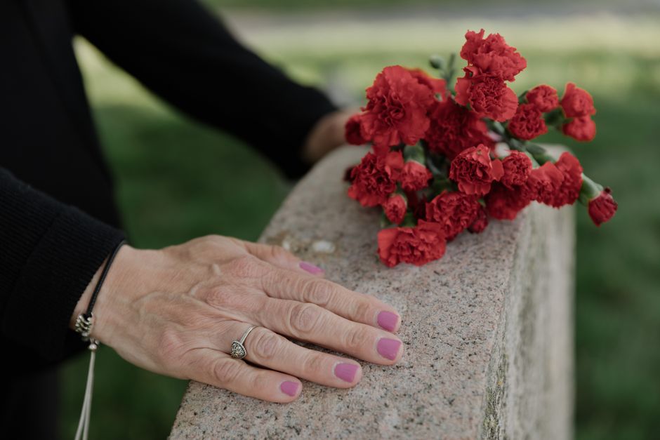 A pair of hands with pink-painted nails rests gently on a stone ledge next to a bouquet of vibrant red carnations, which are arranged in a casual, natural style. The person is wearing a black long-sle