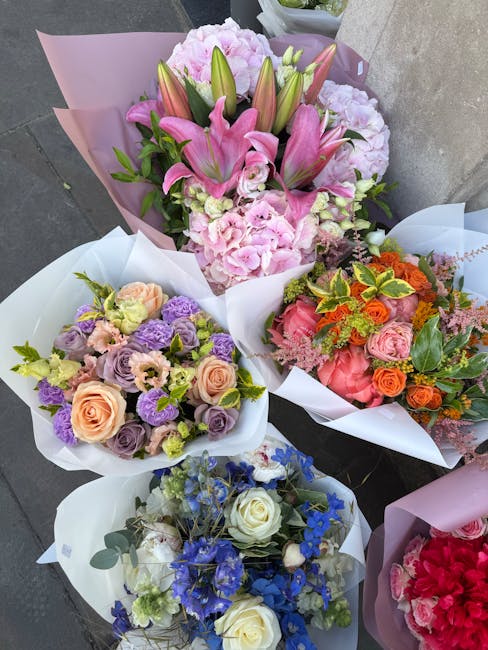 A collection of four floral bouquets displayed on a grey surface. The central bouquet features pink lilies, peonies, and buds with green leaves, wrapped in pink and purple tissue paper. To the left, a