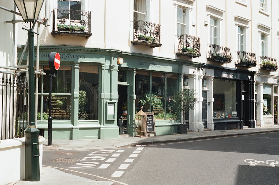 A charming, historic flower shop with a soft pastel green façade featuring large glass windows displaying fresh floral arrangements inside. The shop has decorative iron balconies above, adorned with