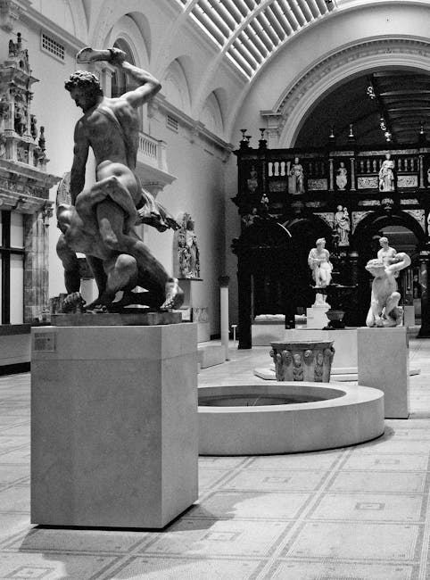 A black and white photograph showing several classical marble sculptures displayed inside a museum with a high, arched glass ceiling. In the foreground, a sculpture depicts a dynamic scene with a figu